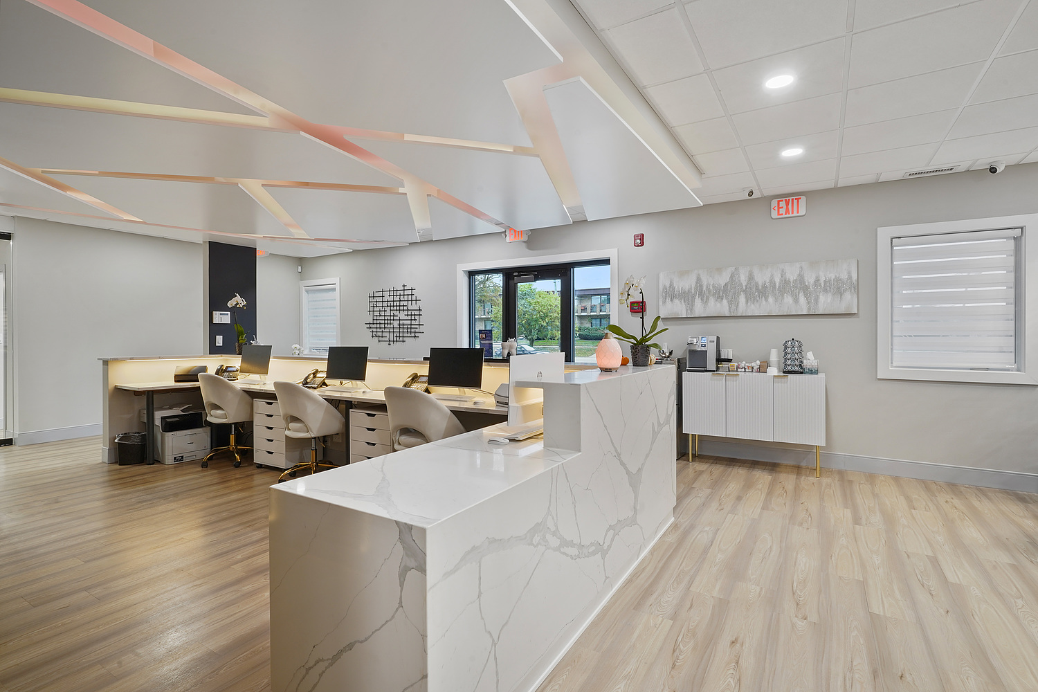 The image shows a modern office space with a clean and minimalist design, featuring a reception area with a desk, chairs, and a computer monitor, a waiting area with a seating arrangement, and an open-concept work environment with desks and cubicles.