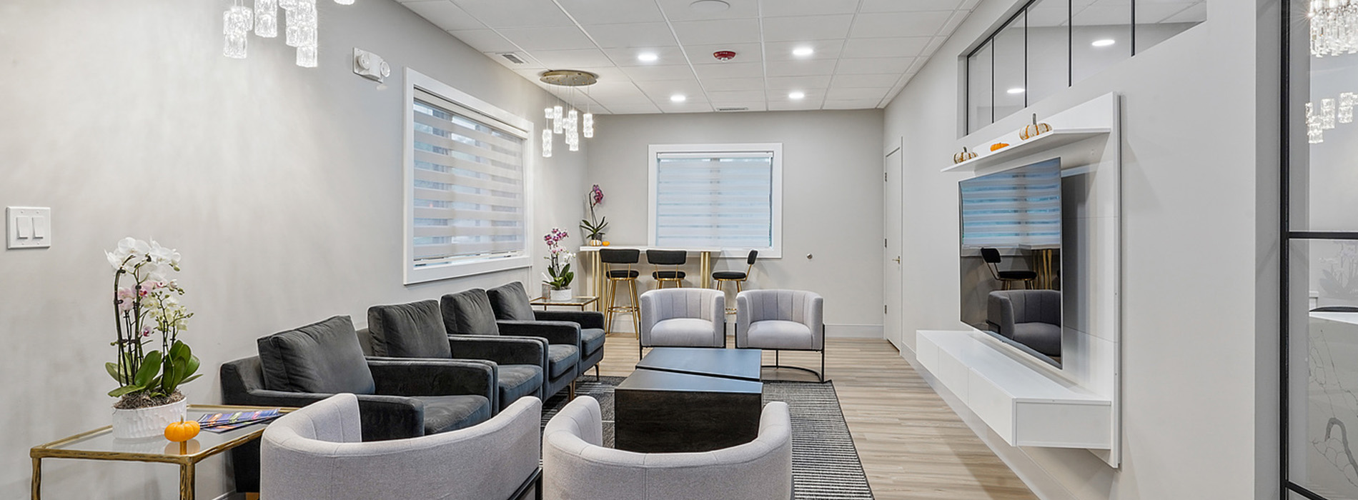 The image shows a modern, well-lit waiting area with contemporary furniture and decor, featuring seating areas, a reception desk, and a large window.