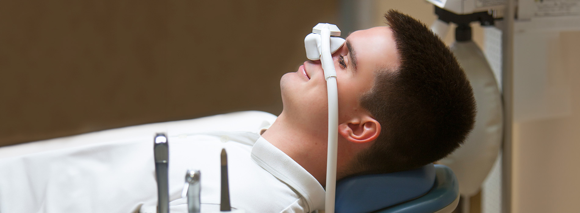 A man sitting in a dental chair with an open mouth, wearing a white shirt and a face mask, looking at the camera.