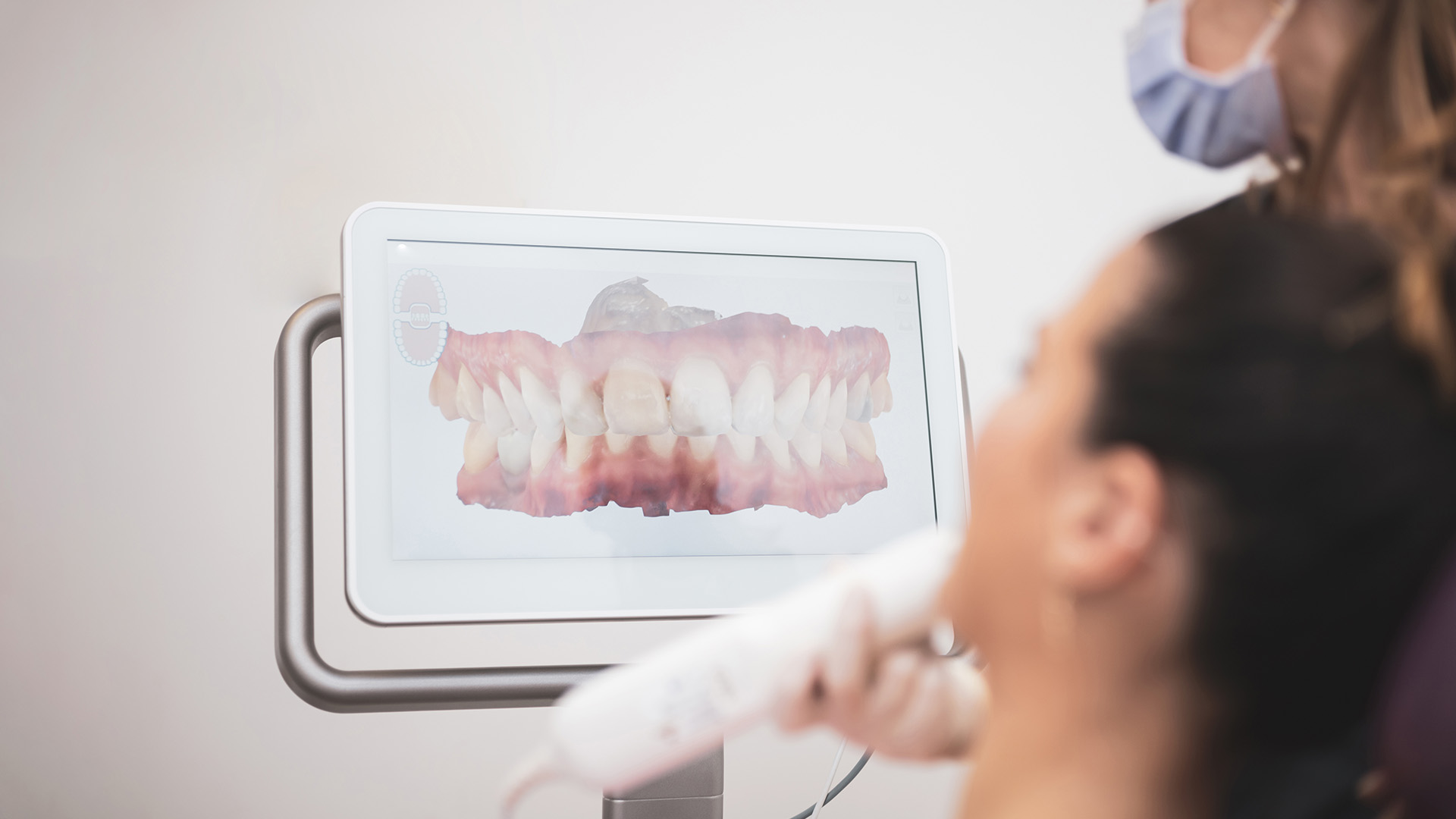 The image shows a person sitting in front of a computer monitor displaying an X-ray, with another individual standing behind the seated person, both appearing to be engaged in a dental or medical setting.