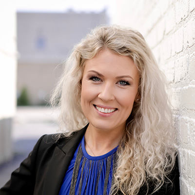 A smiling woman with blonde hair, wearing a blue top and standing against a brick wall.