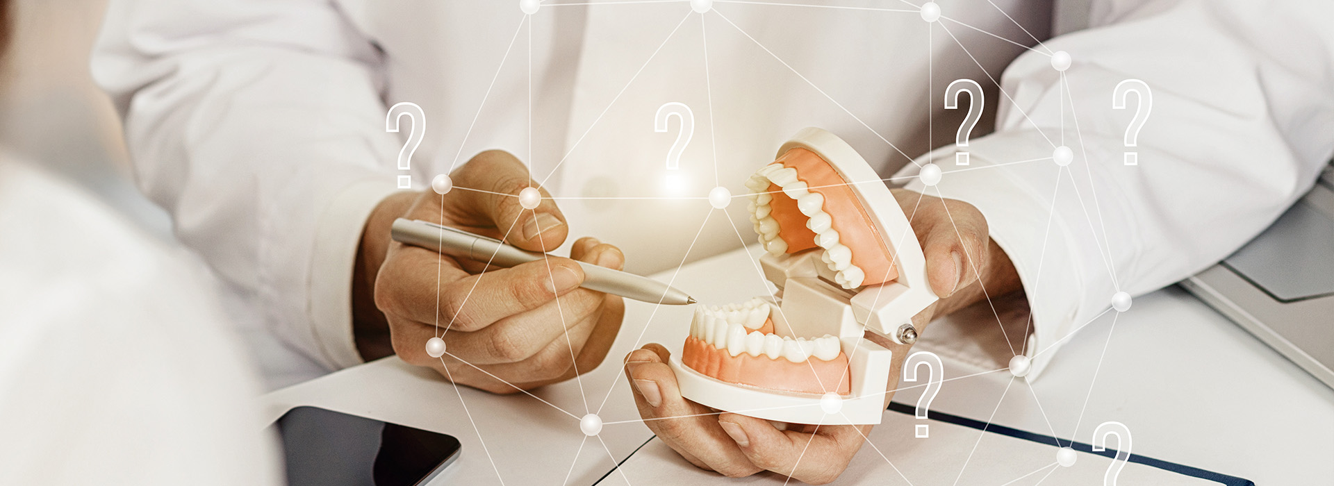 The image shows a person s hand holding a dental implant model, with another individual in the background wearing a white lab coat and examining the model.