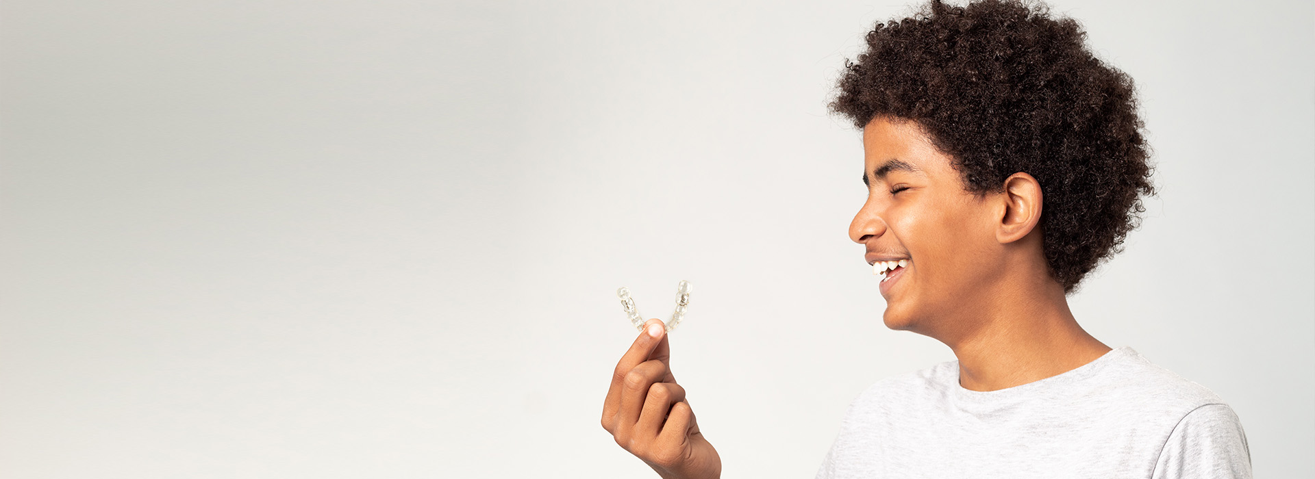 A young person is smiling and holding a small object, possibly a flower or leaf, in front of their face. The background is neutral with a soft gradient from dark to light.