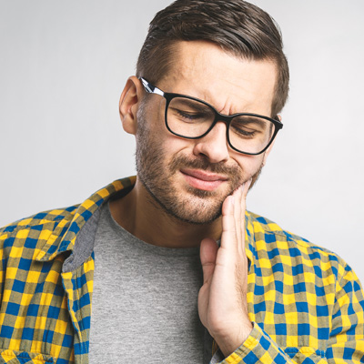 The image depicts a man with glasses, wearing a yellow plaid shirt and holding his hand to his mouth. He appears to be in pain or discomfort, as suggested by his facial expression.