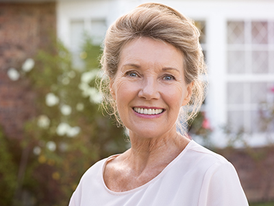 An elderly woman with white hair, wearing a light-colored top and smiling at the camera, standing in front of a brick house with a garden.