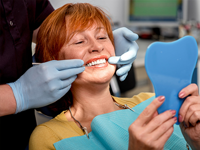 The image shows a woman sitting in a dental chair, smiling as she looks at her reflection in a mirror. She is wearing a blue mouthguard and has white gloves on her hands. A dental hygienist is standing behind her, adjusting the mouthguard with one hand while holding a small blue model of a mouthguard with the other.