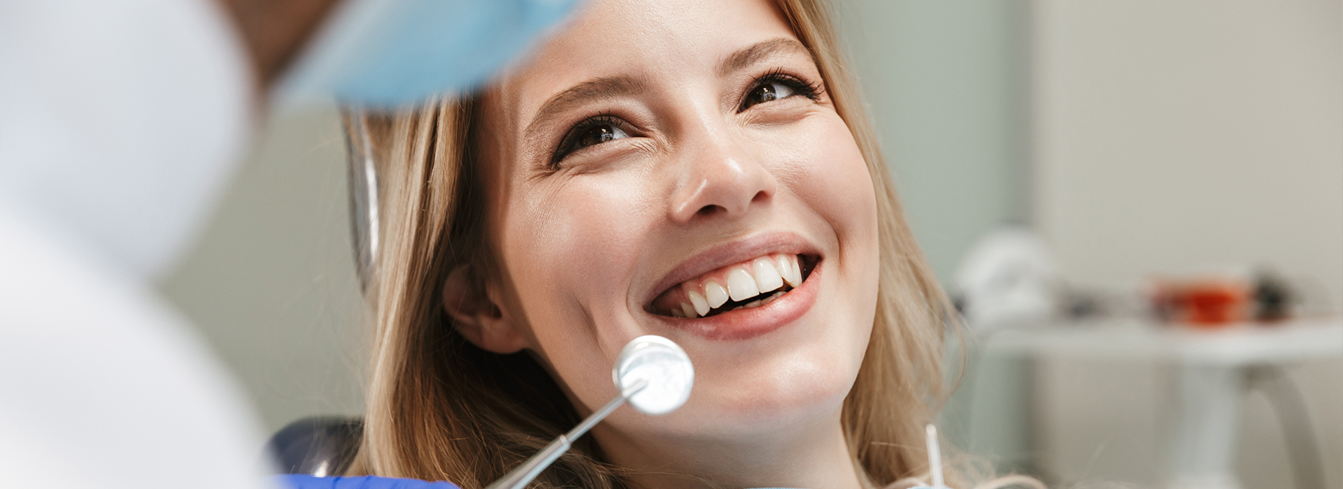 The image shows a woman with a smile, sitting in front of a dental professional who is holding a mirror and observing her teeth.