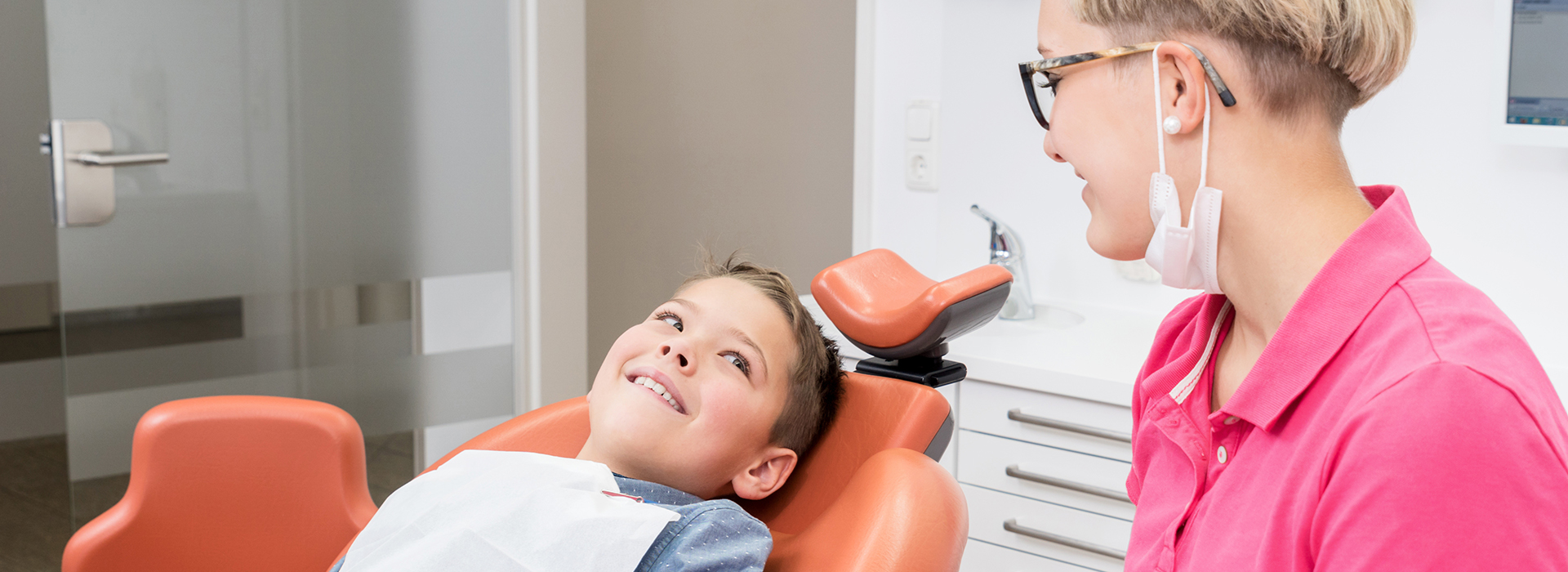 The image shows a dental office setting with a person lying in a dental chair, receiving treatment. A dentist is standing beside the patient, smiling and looking at the camera.