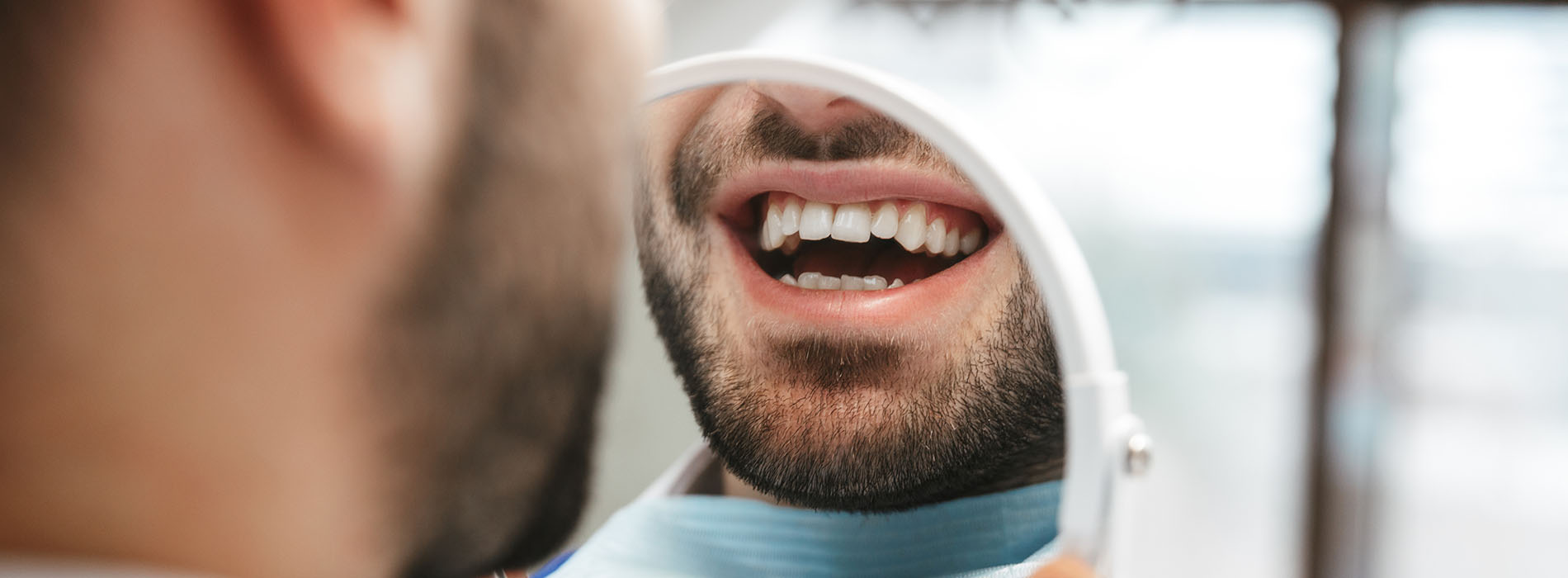 A man in a dental office holding up an object to show his teeth, with another person standing behind him.