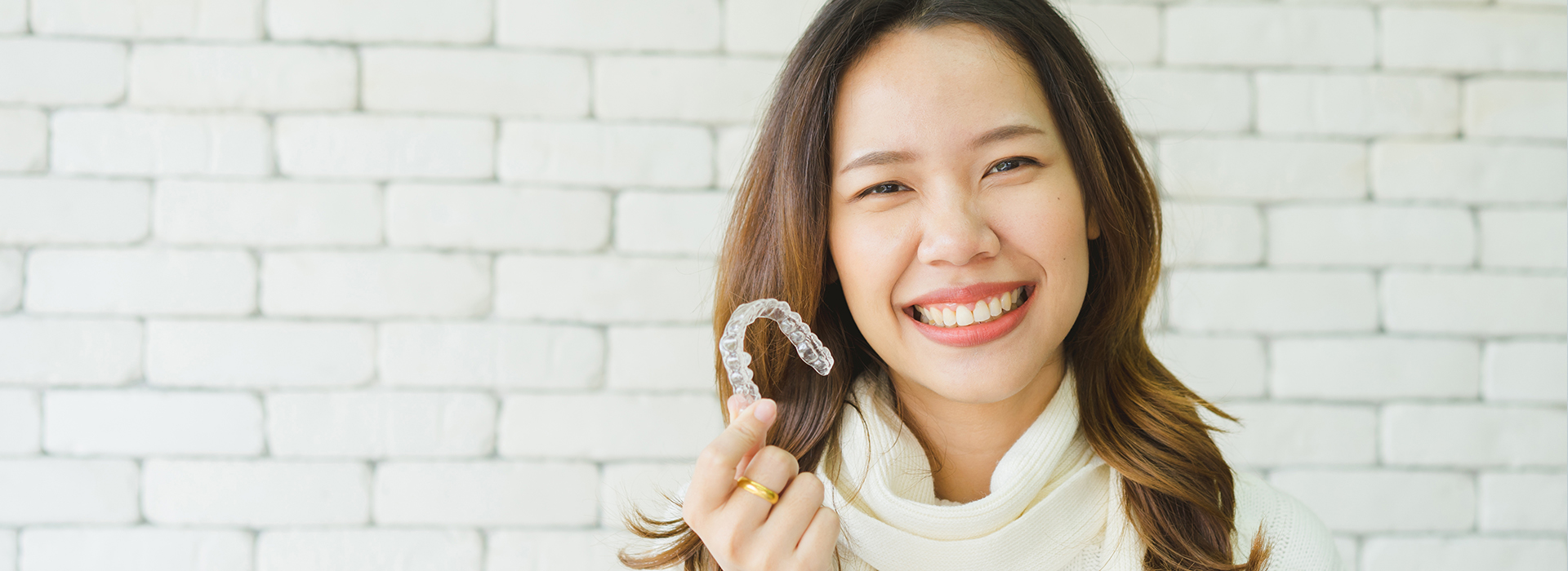 A woman is smiling and holding a ring in her hand, set against a white brick wall.