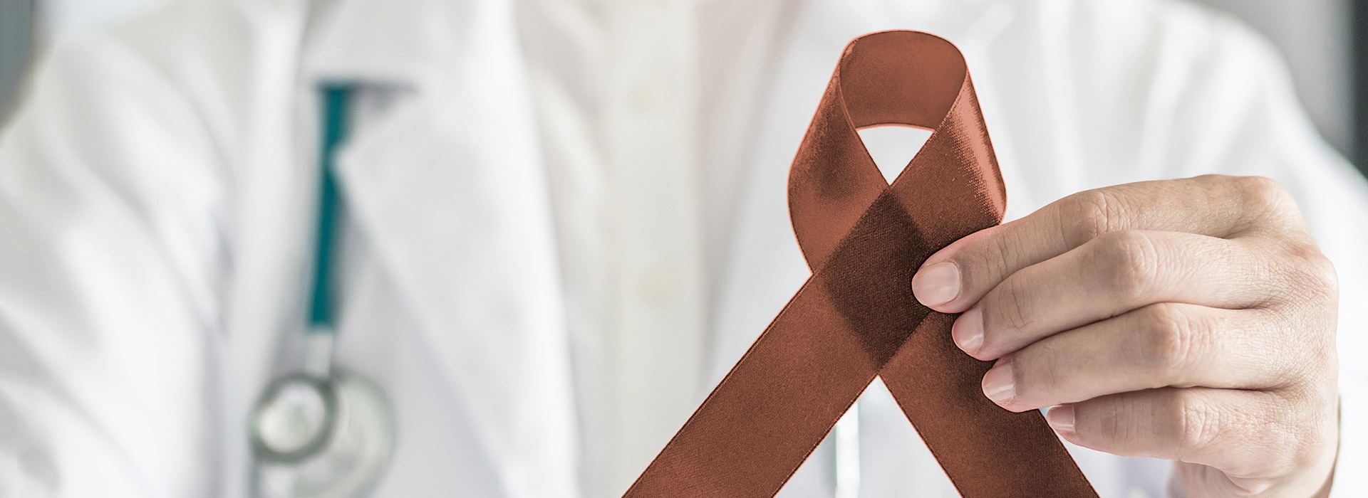 The image shows a medical professional holding up a brown ribbon with a red symbol on it, which is commonly associated with awareness for cancer.