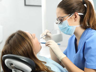 The image shows a dental hygienist performing dental work on a patient, with the patient seated in a dental chair and wearing a surgical mask.