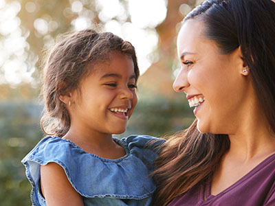 The image is a photograph capturing a joyful moment between a woman and a young child, with the woman smiling at the camera and the child looking towards her with a smile. They appear to be outdoors during daylight.