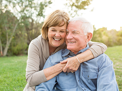 An elderly couple embracing in a park setting, with the woman wearing a light-colored top and glasses, and the man in a blue shirt and dark pants.