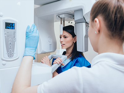 A woman in a blue uniform is operating a large, white 3D scanner while a person in a white lab coat and gloves observes.