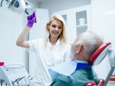 A dental professional in a white coat and purple gloves, smiling at the camera while standing next to an elderly patient with a medical mask covering their face.