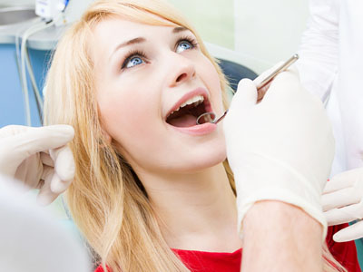 A young woman with blonde hair is sitting in a dental chair, receiving dental care from a professional. She appears to be experiencing some discomfort or pain, as suggested by her facial expression and the presence of dental tools around her mouth.