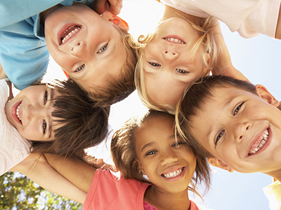 A group of six children, both boys and girls, smiling at the camera in a sunlit outdoor setting with blue sky.