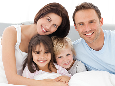 A family of four, including two adults and two children, posing for a photograph in bed with a smile.