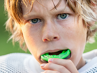 A young boy with blonde hair, wearing a white football jersey, is captured in mid-chew of a green object while holding it with his mouth.