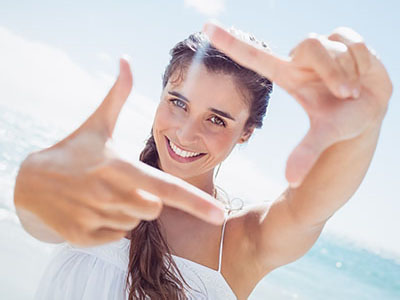 The image shows a woman taking a selfie with her hand, smiling and looking directly at the camera. She is outdoors in bright sunlight, wearing sunglasses, and has long hair.